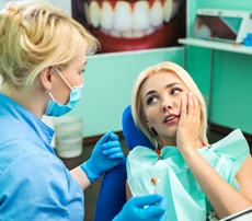 A young woman holding her cheek while her dentist prepares to examine her smile and find the problem area
