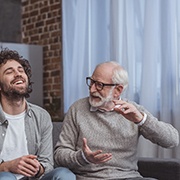Older man speaking to a laughing younger man in front of a curtained window